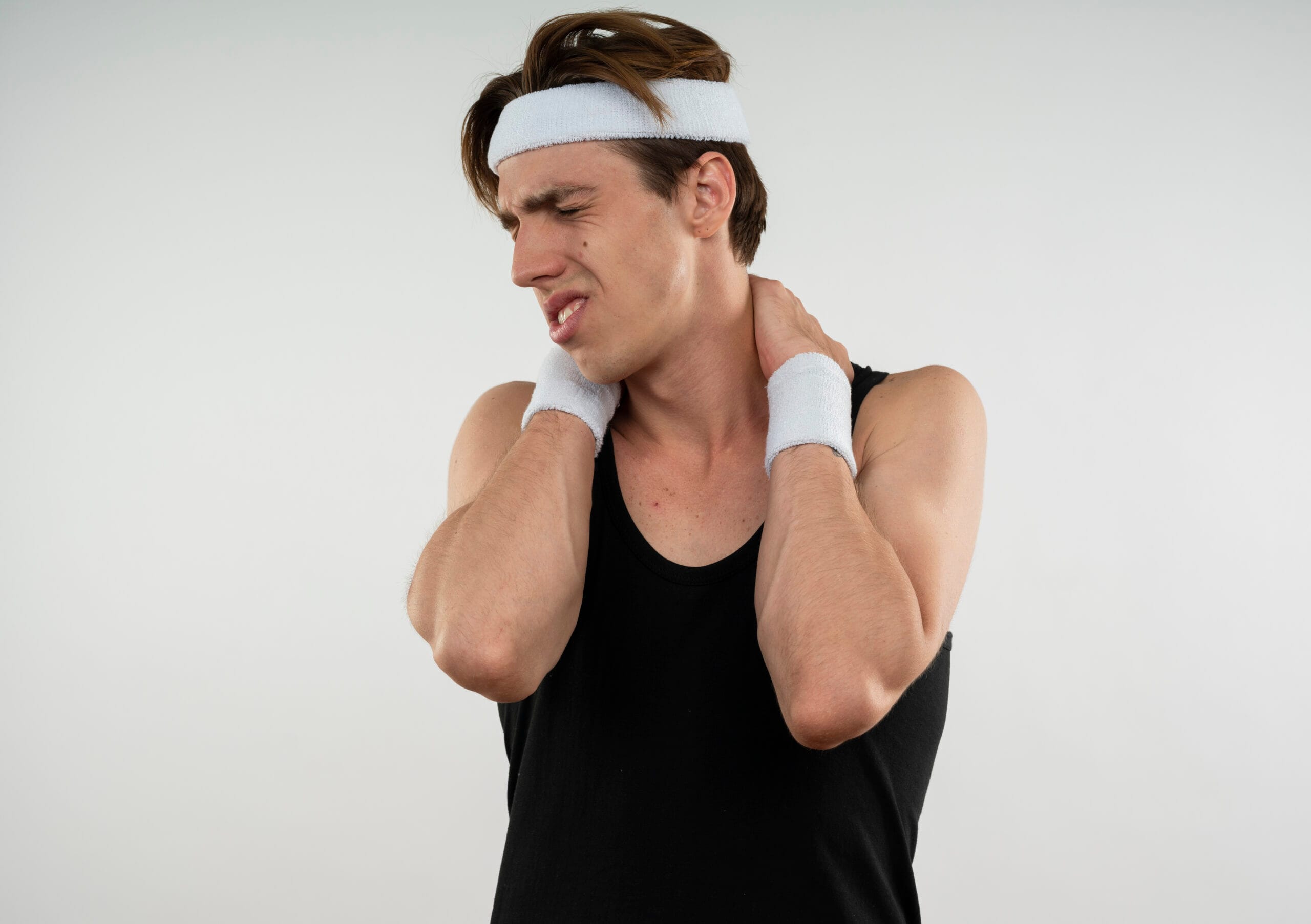 unpleased young sporty guy with closed eyes wearing headband and wristband grabbed neck isolated on white background