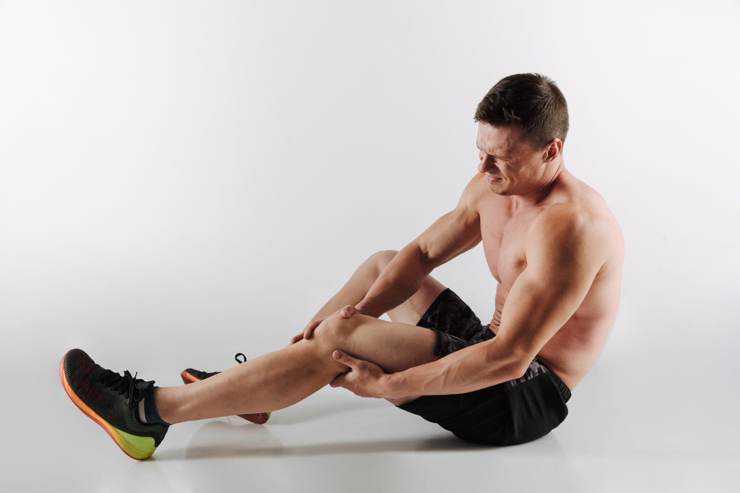 Image of displeased young sportsman standing isolated over grey background. Looking aside have painful feelings in leg.